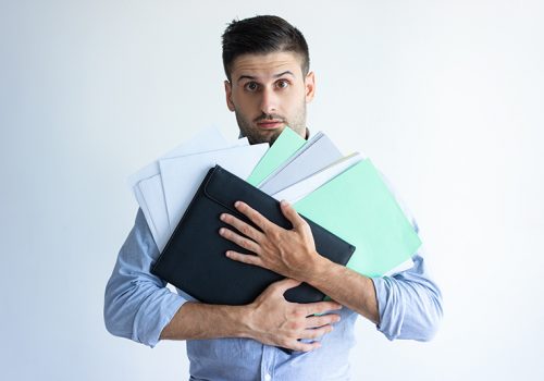 Puzzled office worker holding pile of documents. Young Caucasian man holding heap of papers and folder. Paperwork concept